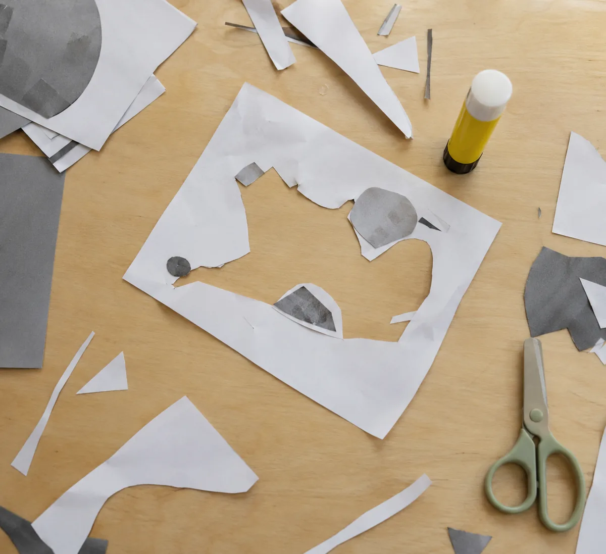 Recortes de papel soltos e sem forma espalhados na mesa, com tesoura sem ponta e cola ao lado. Imagem ilustrativa